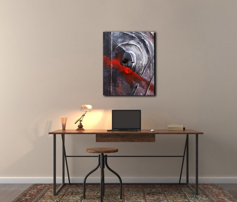 May include: Abstract painting of a face with a red stripe across the eyes. The painting is framed and hanging on a wall above a desk with a lamp, a laptop, and a stool.