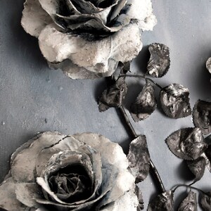 May include: Two white roses with black centers and silver accents are arranged with black leaves on a gray background.