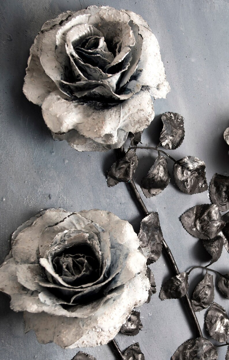 May include: Two white roses with black centers and silver accents are arranged with black leaves on a gray background.