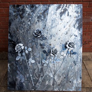 May include: A square abstract painting with raised grey roses and blue-grey leaves and stems. The background is a textured mix of grey, white, and silver paint. The artwork is mounted on a wooden frame, creating a three-dimensional effect.