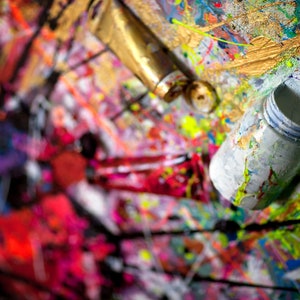 May include: A close-up of a colorful abstract painting with various paint tubes and containers. The paint is splattered and layered, creating a textured and dynamic surface.