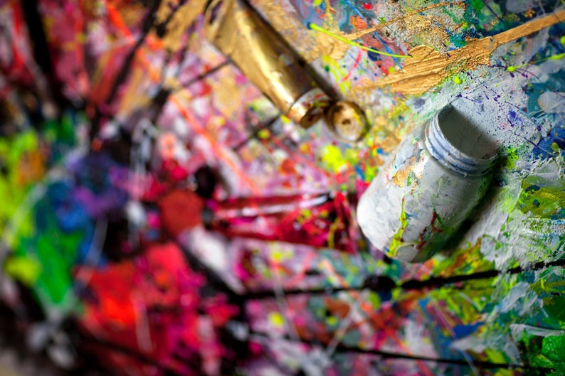 May include: A close-up of a colorful abstract painting with various paint tubes and containers. The paint is splattered and layered, creating a textured and dynamic surface.