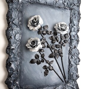 May include: A black and white framed wall art piece featuring three silver roses with black stems and leaves. The frame is textured and has a rough, uneven surface.