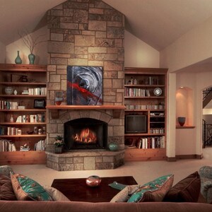 May include: A living room with a stone fireplace, a large window, and built-in bookshelves. The fireplace has a fire burning in it and a painting above it. There are two couches in front of the fireplace, and a coffee table between them.