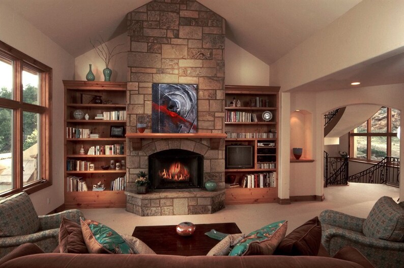 May include: A living room with a stone fireplace, a large window, and built-in bookshelves. The fireplace has a fire burning in it and a painting above it. There are two couches in front of the fireplace, and a coffee table between them.
