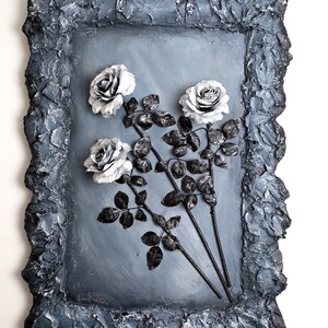 May include: A framed artwork featuring three white roses with black stems and leaves against a gray background. The frame is made of a textured, dark gray material.