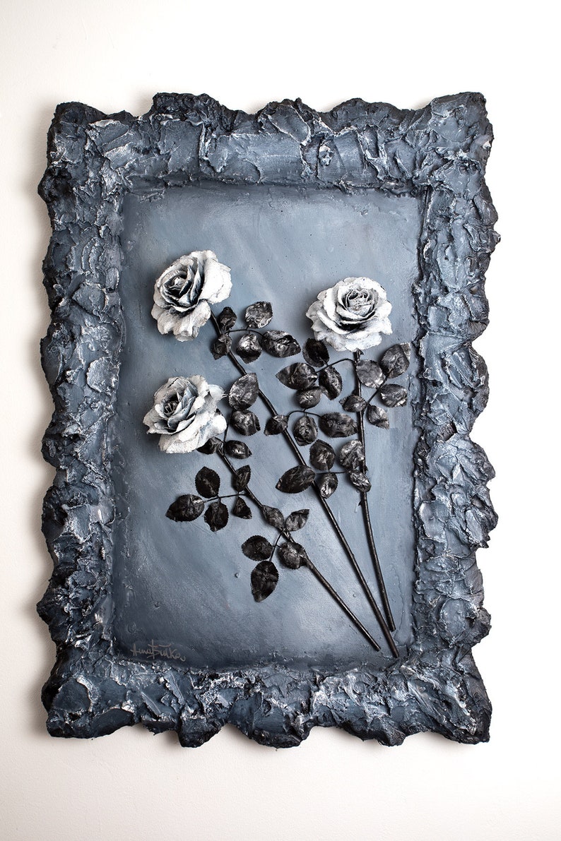 May include: A framed artwork featuring three white roses with black stems and leaves against a gray background. The frame is made of a textured, dark gray material.