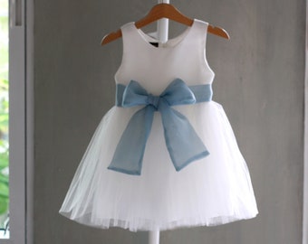 Può includere: Abito di tulle bianco con un fiocco di raso blu. L'abito ha un corpetto senza maniche e una gonna ampia.