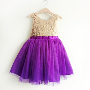 May include: A gold sequin bodice and purple tulle skirt dress for girls. The dress has a purple satin ribbon around the waist.