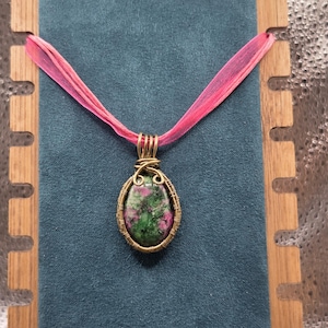 May include: A handcrafted pendant necklace featuring an oval-shaped gemstone with green and pink hues, encased in a gold-toned wire frame. The pendant hangs from a sheer pink ribbon, displayed against a teal backdrop.