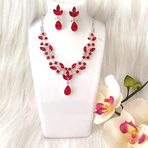 May include: A red and silver necklace and earring set displayed on a white bust. The necklace features a floral design with red gemstones and silver accents. The earrings are teardrop-shaped with red stones.