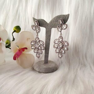 May include: A pair of silver-toned chandelier earrings featuring multiple marquise-cut clear crystals arranged in a leaf-like cluster pattern. The earrings are suspended from a small crystal stud and are displayed on a gray velvet jewelry stand.