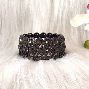 May include: A dark gray stretch bracelet with a repeating pattern of oval-shaped rhinestones. The bracelet is set against a white, fluffy background, creating a contrast that highlights the jewelry's details. The bracelet is a fashion accessory.