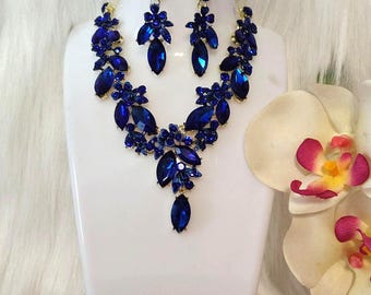 May include: A dazzling blue jewelry set, including a necklace and matching earrings, displayed on a white mannequin. The necklace and earrings feature intricate floral designs with large, faceted blue gemstones. The set is a striking accessory.