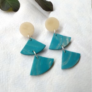 Op de afbeelding: Handgemaakte oorbellen met een crèmekleurige cirkelvormige oorbel en turquoise, waaiervormige hangers. De hangers hebben een marmer effect. De oorbellen zijn verbonden met zilverkleurige ringetjes. De achtergrond is een wit, getextureerd oppervlak.