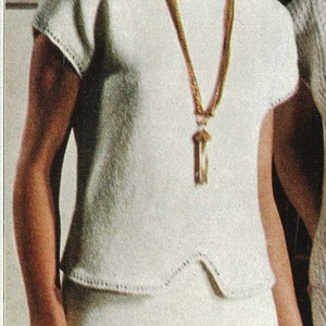 May include: A woman in a white knitted top and skirt, captured in a vintage photograph. The top features short sleeves and a round neckline. A gold necklace with a pendant is worn. The woman has blonde hair. The background includes a painting.