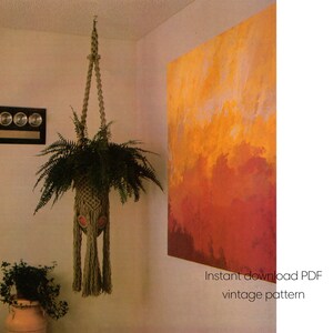 May include: A hanging macrame plant holder with a fern inside. The plant holder is beige and has a decorative pattern with tassels. The image also shows a large abstract painting in shades of orange and red, and the text "Instant download PDF vintage pattern".