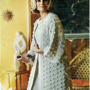 May include: A woman wearing a white knitted cardigan with a textured pattern. The cardigan is long and has a loose fit. The woman is standing in a room with a wooden floor and a yellow wall. The text "BY Villawool" is visible in the upper right corner of the image. The text "4' FIFTH EDITION" is visible in the lower left corner of the image.