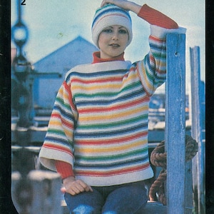 May include: A woman wearing a rainbow striped sweater with a white crocheted hat and blue jeans. She is sitting on a wooden bench with a rope tied to the side.