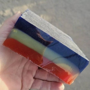 May include: A multi-colored layered soap bar with a dark blue, light green, and red color scheme. The soap bar is translucent and has a glossy finish.