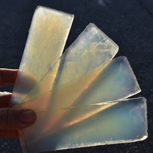 May include: Four translucent, rectangular opalescent stones are fanned out, catching the sunlight. The stones have a milky, iridescent quality with hints of yellow and orange. The edges are rough, and the overall effect is ethereal.