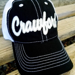 May include: Black and white trucker hat with a white mesh back and a white embroidered "Crawford" on the front. The hat has a black brim with white stitching.