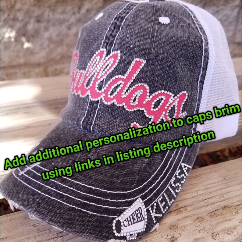May include: A black and white distressed baseball cap with a pink rhinestone design that says "Bulldogs". The cap has a white mesh back and a white stitched brim. The word "CHEER" is on the side of the cap with rhinestones. The name "KELLISSA" is written on the side of the cap.