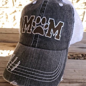 May include: A black and white distressed baseball cap with a brown paw print and the word "MOM" in rhinestones.