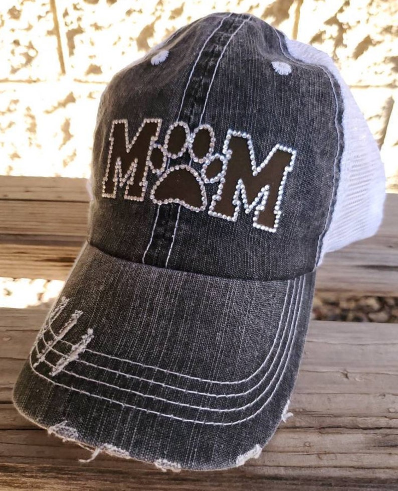 May include: A black and white distressed baseball cap with a brown paw print and the word "MOM" in rhinestones.