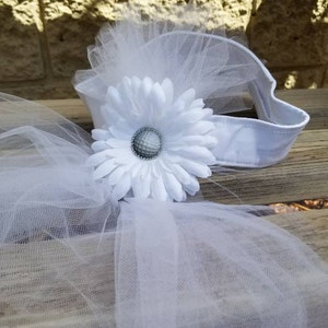 K&ouml;nnte beinhalten: Ein wei&szlig;es T&uuml;ll-Haarband mit einer wei&szlig;en G&auml;nsebl&uuml;mchen-Blume und einem silbernen Golfball in der Mitte. Das Haarband liegt auf einer Holzoberfl&auml;che.