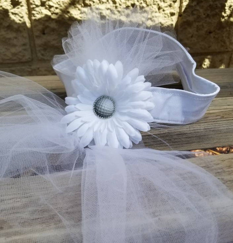 K&ouml;nnte beinhalten: Ein wei&szlig;es T&uuml;ll-Haarband mit einer wei&szlig;en G&auml;nsebl&uuml;mchen-Blume und einem silbernen Golfball in der Mitte. Das Haarband liegt auf einer Holzoberfl&auml;che.
