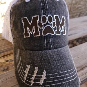 May include: A black and white distressed baseball cap with a brown patch that says "MOM" with a paw print in the middle. The cap has a white mesh back.