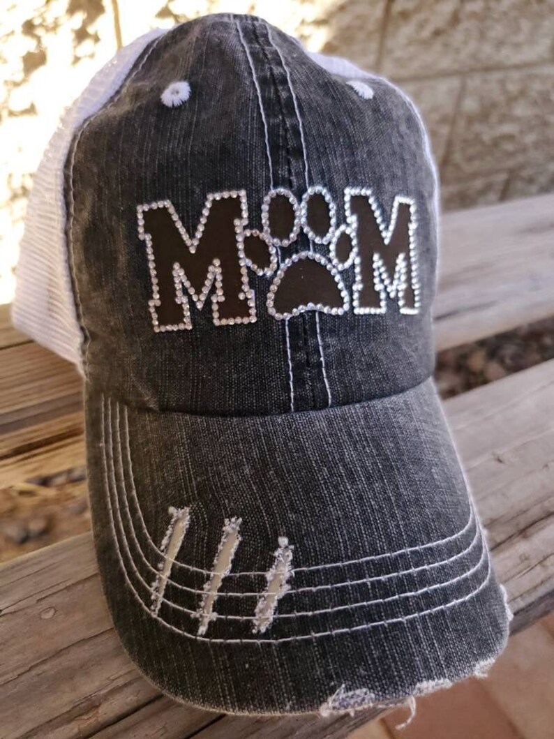 May include: A black and white distressed baseball cap with a brown patch that says "MOM" with a paw print in the middle. The cap has a white mesh back.
