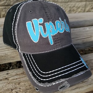 May include: A gray and black baseball cap with a distressed look. The front of the cap features the word "VIPERS" in turquoise with rhinestones. The brim of the cap is black with white stitching.