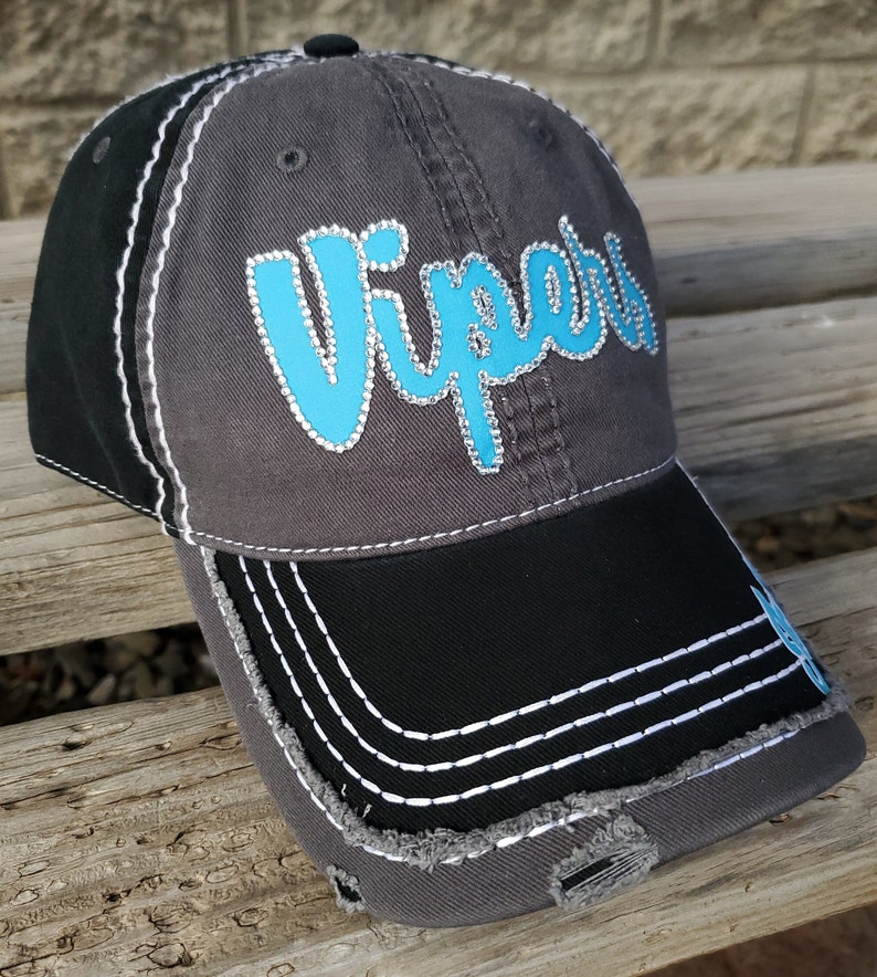 May include: A gray and black baseball cap with a distressed look. The front of the cap features the word "VIPERS" in turquoise with rhinestones. The brim of the cap is black with white stitching.