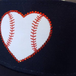 May include: A navy blue baseball cap featuring a white heart-shaped graphic with red baseball stitching details. The heart design is outlined with small, sparkling red rhinestones that follow the perimeter of the shape on the front panel.