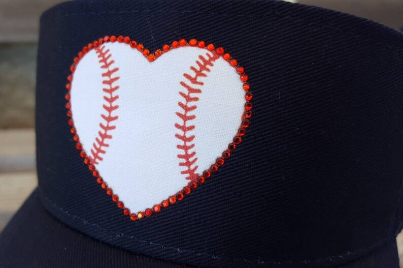 May include: A navy blue baseball cap featuring a white heart-shaped graphic with red baseball stitching details. The heart design is outlined with small, sparkling red rhinestones that follow the perimeter of the shape on the front panel.