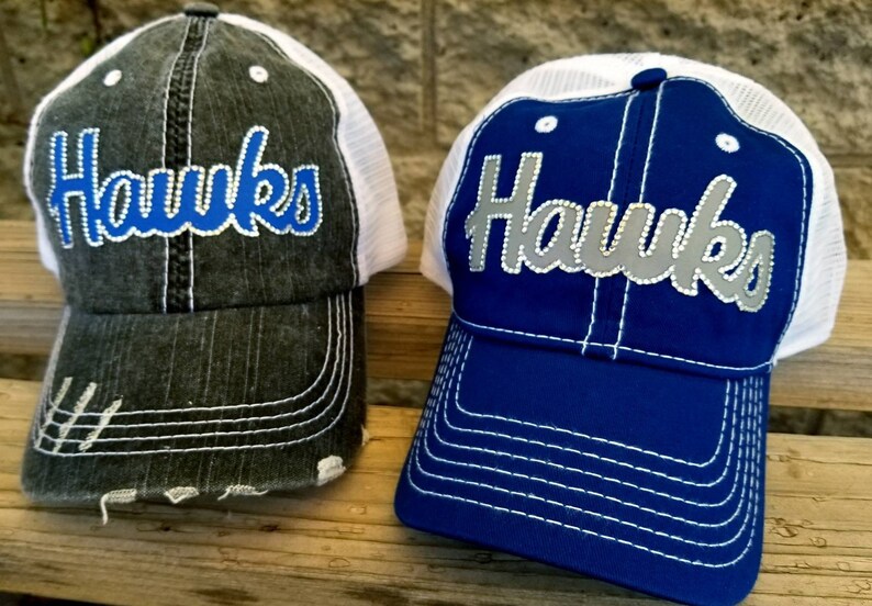 Puede incluir: Dos gorras de camionero con la palabra "Hawks" bordada en la parte delantera. Una gorra es negra con malla blanca y letras azules. La otra gorra es azul con malla blanca y letras plateadas.