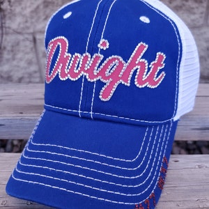 May include: A blue and white trucker hat with a red and white "Dwight" design with rhinestones. The hat has a mesh back and a white brim.