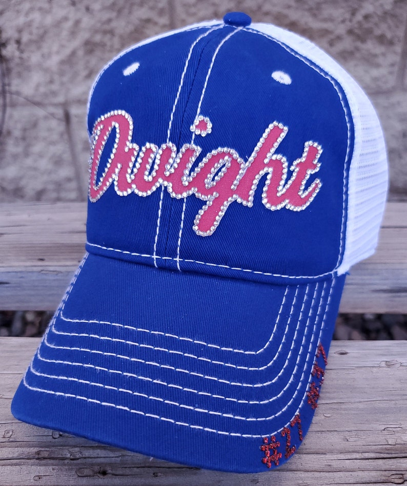 May include: A blue and white trucker hat with a red and white "Dwight" design with rhinestones. The hat has a mesh back and a white brim.
