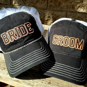 May include: Two black trucker hats with white mesh back panels. The hat on the left has a white veil and says "BRIDE" in orange with a rhinestone trim. The hat on the right says "GROOM" in orange.