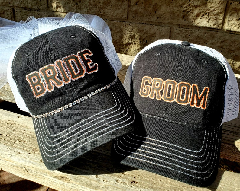 May include: Two black trucker hats with white mesh back panels. The hat on the left has a white veil and says "BRIDE" in orange with a rhinestone trim. The hat on the right says "GROOM" in orange.