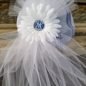 May include: A white tulle veil with a white flower and a blue and white New York Yankees logo pin. The pin has rhinestones around the edge.