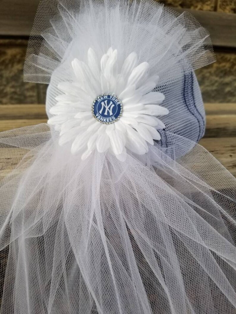 May include: A white tulle veil with a white flower and a blue and white New York Yankees logo pin. The pin has rhinestones around the edge.