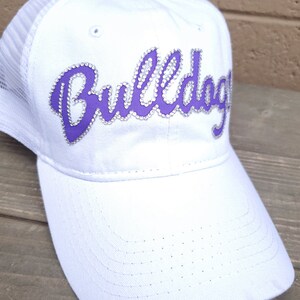 Puede incluir: Gorra de camionero blanca de malla con un logotipo "Bulldogs" morado con pedrer&iacute;a.