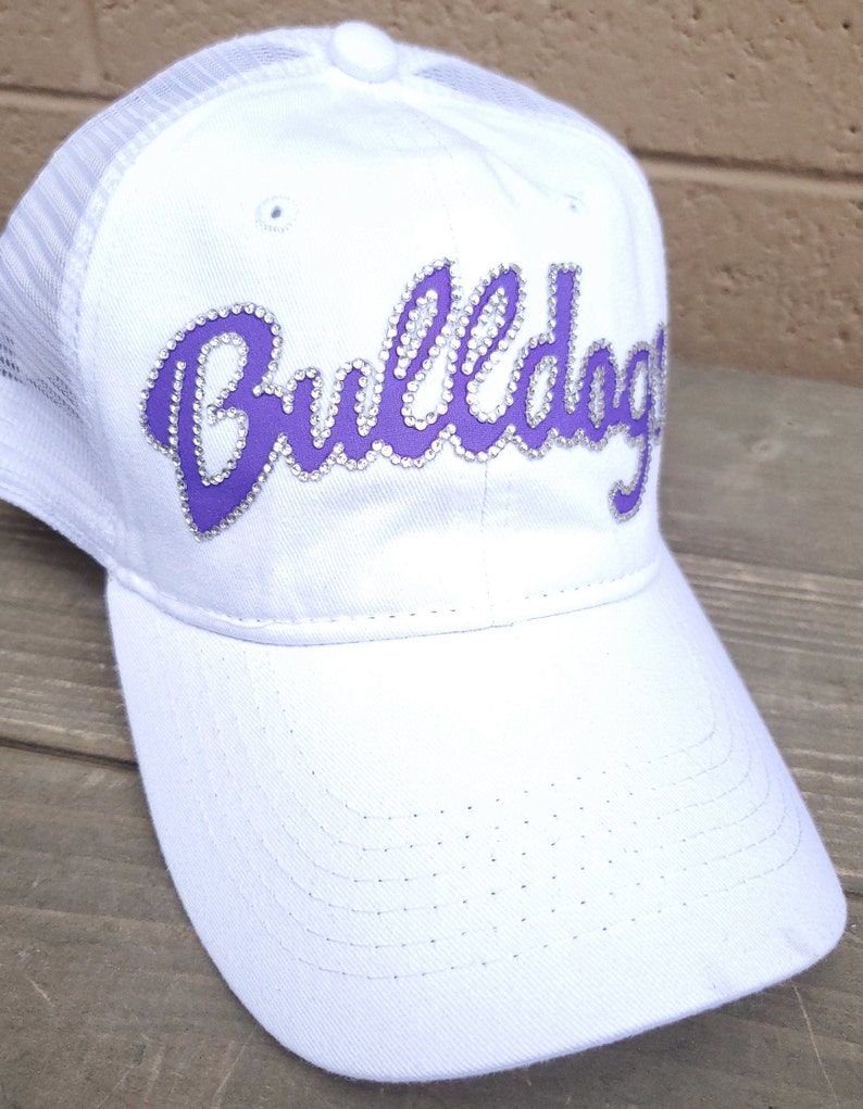 Puede incluir: Gorra de camionero blanca de malla con un logotipo "Bulldogs" morado con pedrer&iacute;a.