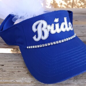 K&ouml;nnte beinhalten: Ein k&ouml;nigsblauer Visier mit einem wei&szlig;en "Bride"-Design, das mit Strasssteinen verziert ist. Das Visier hat einen wei&szlig;en T&uuml;llschleier, der oben befestigt ist.