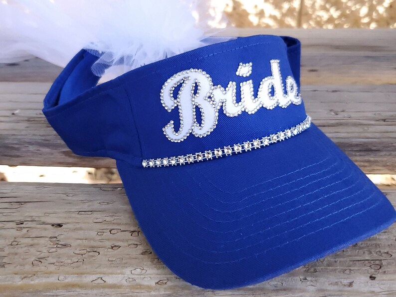 K&ouml;nnte beinhalten: Ein k&ouml;nigsblauer Visier mit einem wei&szlig;en "Bride"-Design, das mit Strasssteinen verziert ist. Das Visier hat einen wei&szlig;en T&uuml;llschleier, der oben befestigt ist.
