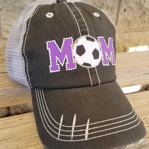 Op de afbeelding: Een zwarte en grijze baseballpet met een mesh achterkant. De voorkant van de pet heeft een paarse "MOM" met een witte voetbal in het midden.