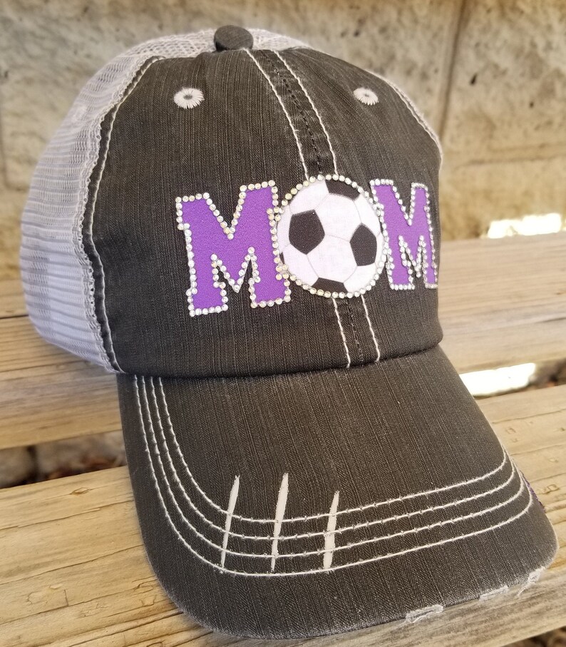 Op de afbeelding: Een zwarte en grijze baseballpet met een mesh achterkant. De voorkant van de pet heeft een paarse "MOM" met een witte voetbal in het midden.